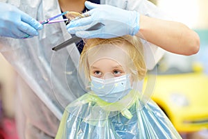 Preschooler boy wearing facemask getting haircut while coronavirus epidemic. Children hairdresser with professional tools - comb