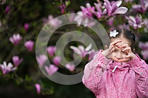 Preschooler girl in pink jacket showing heart by fingers while enjoying nice spring day near magnolia blooming tree