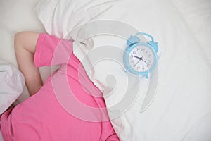 a preschooler girl in a pink t-shirt hides her head under a pillow, an alarm clock on the bed