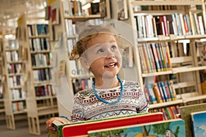 Preschooler little girl sitting and reading a book in library. Kid with books near a bookcase. Happy, cheerful and cute girl