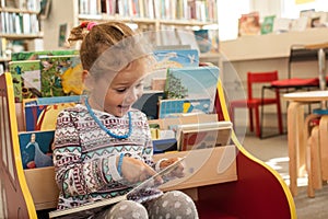 Preschooler little girl sitting and reading a book in library. Kid with books near a bookcase. Happy, cheerful and cute girl read