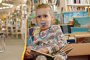 Preschooler little girl sitting and reading a book in library. Kid with books near a bookcase. Happy, cheerful and cute girl read