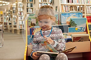 Preschooler little girl sitting and reading a book in library. Kid with books near a bookcase. Happy, cheerful and cute girl read