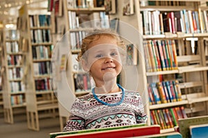 Preschooler little girl sitting and reading a book in library. Kid with books near a bookcase. Happy, cheerful and cute girl read