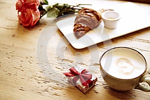 Present box, rose flowers, fresh croissant, coffee on wooden table. Romantic breakfast for Valentine`s Day celebrate.