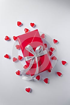 Present box is wrapped in red paper with silver ribbon bow. White background with a pile of red jelly hearts. Top view.