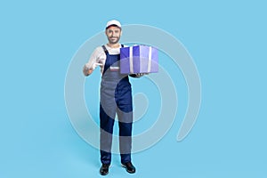 This present for you! Full length happy courier in overalls and protective gloves pointing at big wrapped box
