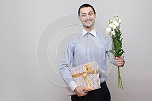 Presents for birthday. Charming pleasure romantic young man in blue shirt holding bouquet of white flowers and gift with yellow bo