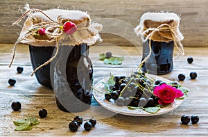 Preserved homemade black currant jam in glass jars on light wooden table. Fresh berries and green leaves, white vintage plate