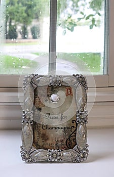 Press for Champagne sign with button to push in ornate silver frame sits in front of wood framed window with screen on rainy summe