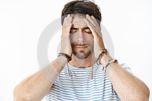 Pressured uneasy male entrepreneur with beard in striped t-shirt having problems at work holding hands on face while