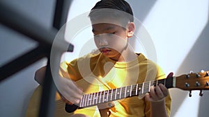 Preteen Asian boy playing acoustic guitar dressed sitting on the cozy sofa at the home living room and enjoying a favorite hobby,