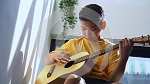 Preteen Asian boy playing acoustic guitar dressed sitting on the cozy sofa at the home living room and enjoying a favorite hobby,