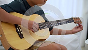 Preteen Asian boy playing acoustic guitar dressed sitting on the cozy sofa at the home living room and enjoying a favorite hobby,