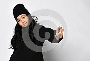 Preteen asian girl in black sportswear and hat showing stop gesture to camera