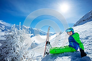 Preteen boy with alpine ski sit in snow over mountain peaks