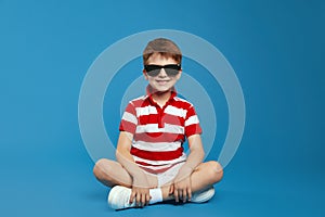 Preteen boy in casual outfit and trendy sunglasses feels happy sitting against the blue background