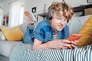 Preteen boy lying at home on cozy sofa dressed casual jeans and shirt. listening to music and chatting using wireless headphones