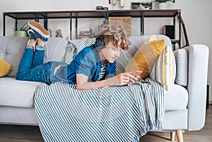 Preteen boy lying at home on cozy sofa dressed casual jeans and shirt. listening to music and chatting using wireless headphones