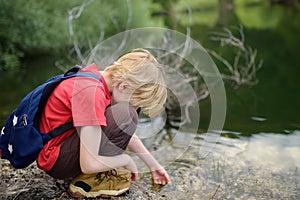 Preteen boy in red shirt is exploring nature and playing with water in lake during hiking in mountains valley. Active leisure for