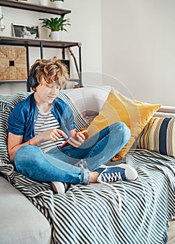 Preteen boy sitting at home on cozy sofa dressed casual jeans and new sneakers listening to music and chatting using wireless