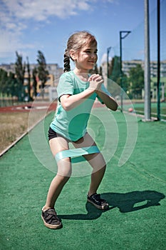 Preteen girl making exercises with fitness resistance band at public sportsground in city, sport