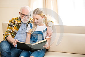 Preteen girl with senior man in eyeglasses reading book together