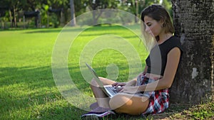 Pretty adult girl is sitting under a tree in the park, keeps the computer on her knees,