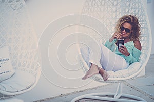 Pretty adult woman sit down on a white comfy seat in outdoor garden smiling and using cell phone to video call - people luxury