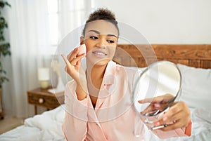 Pretty african american lady applying cosmetic foundation with applicator, holding mirror, sitting on bed in bedroom
