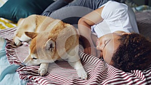 Pretty African American teenage girl and her loyal dog are sleeping together on bed at home, person is hugging animal in