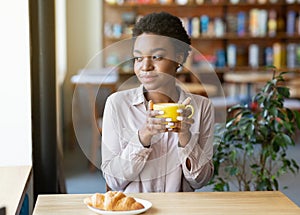 Pretty African American woman having breakfast, enjoying her morning coffee with croissant at city cafe