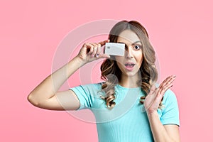 Pretty amazed female holding credit card at her face closing eyes isolated over pink background