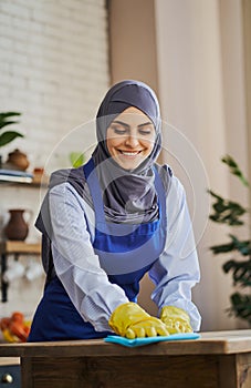 Pretty Arabian woman in apron and gloves cleaning a table in the kitchen