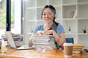 A pretty Asian female college student sits at her study table with a stack of books and her laptop