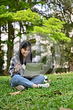 Pretty Asian female college student using her laptop and sitting on grass in the campus park