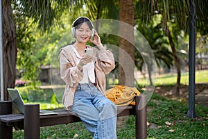 A pretty Asian female student is relaxing on a bench in a park, listening to music on her headphones