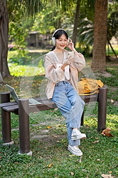 A pretty Asian female student is relaxing on a bench in a park, listening to music on her headphones