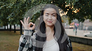 Pretty Asian student showing OK hand gesture outdoors in urban park smiling