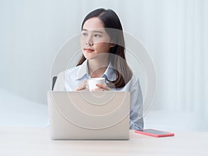 Pretty asian woman hold coffee mug sit in front of laptop computer looking out window thoughtfulness.