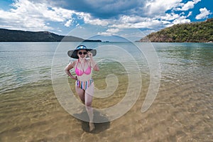 A pretty asian woman in a large sun hat and high waisted bikini enjoys the waters of a golden sand beach at a tropical cove