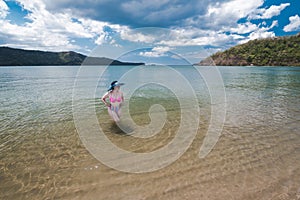 A pretty asian woman in a large sun hat and high waisted bikini takes a dip in the waters of a golden sand beach at a tropical