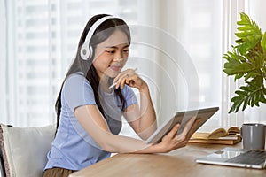 A pretty Asian woman listening to music through her headphones while using her tablet at a table