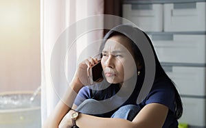 Pretty Asian woman sit and hugging on knees at home using cell phone looking hopelessly at window