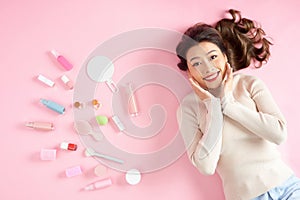 Pretty Asian woman smiling while lying on pink floor with her cosmetic makeup tools. Top view