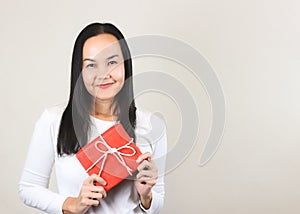 Pretty Asian woman in white long sleeve  t-shirt holding red gift box, smiling and  looking at camera, front view on white