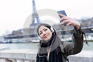 Pretty asian young woman taking a selfie in front of Eiffel tower in Paris while listening to music.