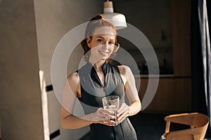 Pretty athletic young woman in sportswear standing by window and drinking fresh water in morning after fitness training