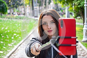 Pretty attractive brunette with amber orange eyes in a public park taking a selfie with a selfie stick and a red smartphone