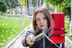 Pretty attractive brunette with amber orange eyes in a public park taking a selfie with a selfie stick and a red smartphone